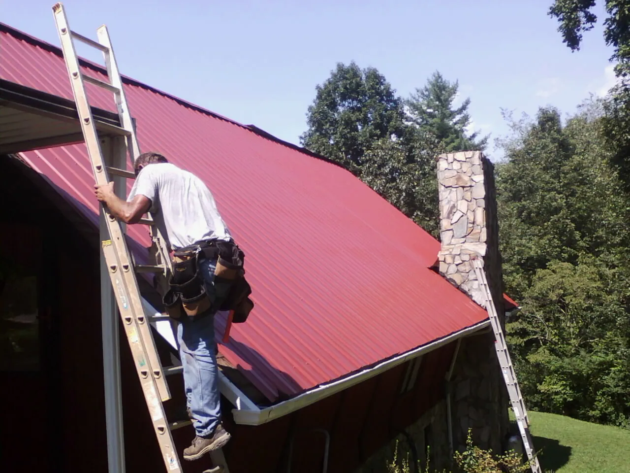 Roofer on a ladder beside a red metal roof and stone chimney — Soffit & Fascia Repair by Roofing Brothers in Lyle